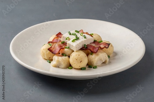 Plate of Bryndzove Halusky topped with crispy bacon, green onions, and feta cheese. Concept of delicious potato dumplings with sheep cheese bacon in close-up.