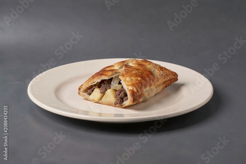 Close-up of Cornish Pasty with golden-brown crust, filling of minced meat, potatoes, swede, onions. Concept of pastry with beef potato swede onion on smooth plate.