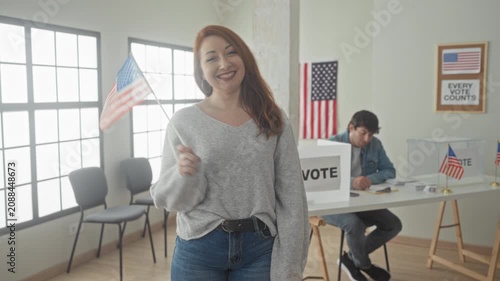 Woman holds small us flag and smiles with hand on hip at a polling station inside a building with vote box and american flags visible; pride civic duty.