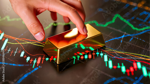 A close-up perspective showing a hand carefully adjusting or pointing at a solid gold bullion bar positioned on a glowing dark surface with complex financial market candlestick charts. , 