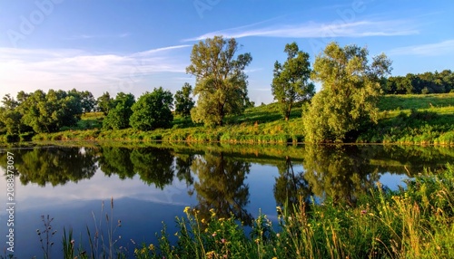 Tranquil river scene with trees reflecting on water under a blue sky with green landscape