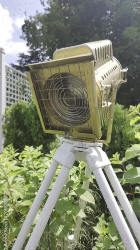 A vintage gold movie spotlight on a white tripod stands outdoors among green bushes. A blue sky with white clouds visible behind the cinematic lighting equipment.
