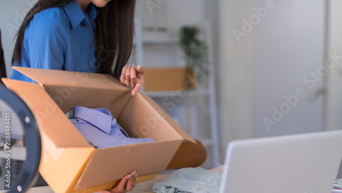 Young Asian woman unpacking cardboard box while managing online merchant business at home office with laptop