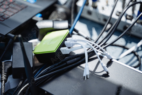 Closeup of portable power bank charging hub with multiple USB cables plugged in, tangled black power leads, adapters, and connectors resting on table in sunlight