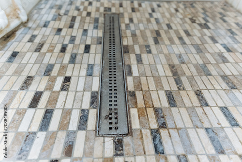 Linear metal shower drain set into beige, cream, brown, and gray mosaic stone tile floor with grout lines, low angle view showing texture, drainage channel, and slight surface debris