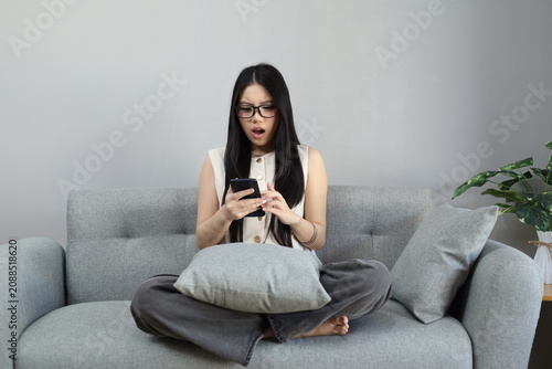 Fotografía A young woman sitting on the sofa at home holding and looking at the phone screen with a shocked expression screaming open her mouth, got the unexpected news