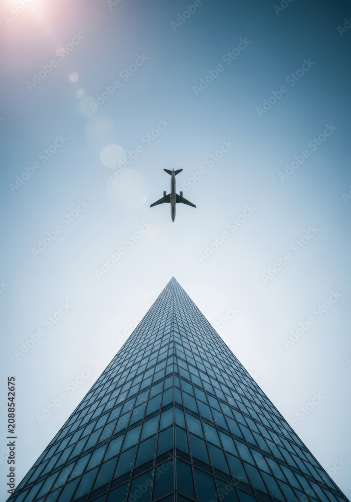 Fototapeta premium Low Angle View of Modern Glass Skyscraper and Commercial Airplane Flying in Clear Blue Sky with Sun Lens Flare