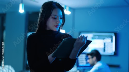 Focused Asian businesswoman uses a smart digital tablet in a modern, blue-lit high-tech corporate office. The female IT professional analyzes secure network data with a colleague in the background.