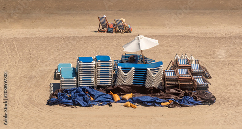 Weymouth Dorset England UK. 02.07.2026. A collection of folded beach loungers and striped deck chairs are neatly piled on a sandy shore, some covered with blue tarps and weighted down
