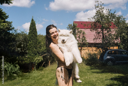 Smiling young woman in bikini holds large fluffy white Samoyed dog in sunny backyard with green lawn, trees and bright blue sky, joyful candid outdoor portrait, with woman, smile