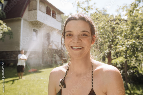 A young woman in a swimsuit cools off in the heat under the spray of water against the backdrop of nature and a country house, backyard, sunny day. A friend is spraying her with a hose.