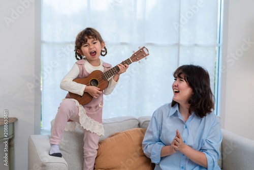 Child plays ukulele while mother smiles in living room during daytime