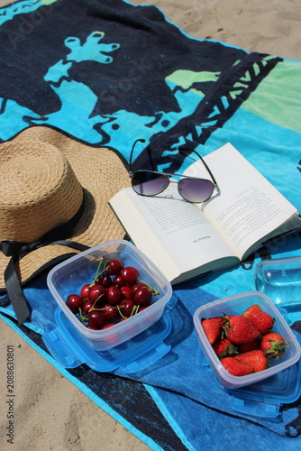 Straw hat and sunglasses rest on an open book placed on a blue beach towel, with containers of cherries and strawberries beside a water bottle on sand