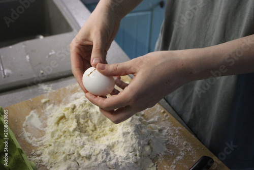 Person's hands crack a white egg above a wooden cutting board with flour, surrounded by kitchen utensils and a sink in the background