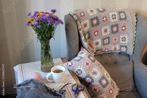 Cozy living room corner with gray armchair, colorful knitted blanket, flowers in vase, cup of tea, yarn, scissors, and crochet project on wooden table