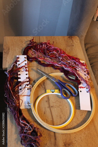 Embroidery materials such as vibrant threads, scissors, and a wooden hoop are neatly placed on a wooden table with a soft fabric backdrop