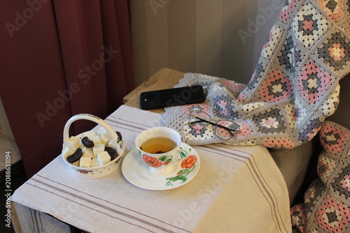 Tea cup with floral design placed on a table beside a basket of sweets and reading glasses, draped with a colorful crochet blanket in a cozy indoor setting