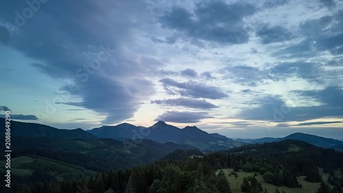 Panoramic mountain dusk landscape with jagged central peak silhouetted against glowing pastel sky, layered cloudbank, forested hills, rolling meadows, and distant ridgelines