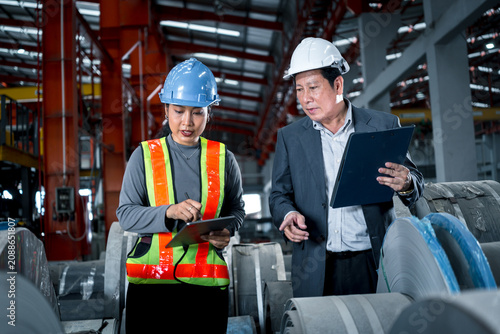 two professional engineer, worker, technician use clipboard discuss work, walk in steel metal manufacture factory plant industry. engineering man and woman wear hard hat check quality machine