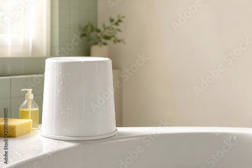 White plastic bucket with water droplets sits on a bathtub edge beside a yellow sponge and soap dispenser in a bright, clean bathroom.