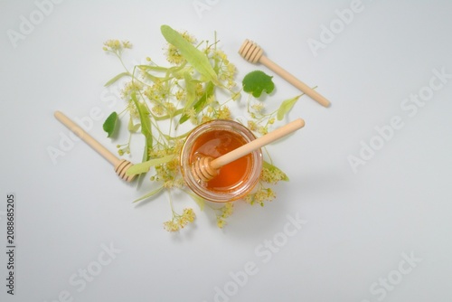 Top view of natural linden honey in a glass jar surrounded by blooming linden flowers with wooden honey dippers on a white background.