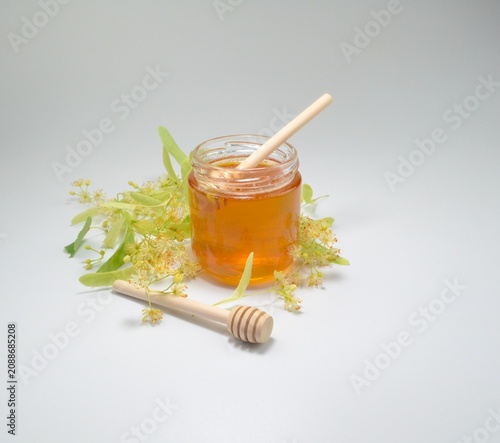 Fresh linden honey in a glass jar decorated with blooming linden flowers and a wooden honey dipper on a clean white background.