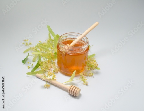 Natural linden honey in a glass jar with a wooden honey dipper surrounded by fresh blooming linden flowers on a white background with copy space.