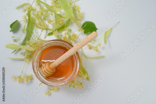 Top view of a glass jar filled with natural linden honey and a wooden honey dipper surrounded by fresh linden blossoms on a white background.