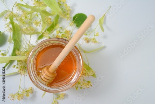 Close-up of natural linden honey in a glass jar with a wooden honey dipper and blooming linden flowers on a white background.