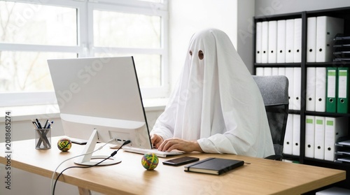 Creative ghostwriter wearing a white sheet costume sitting at a desk with a computer.