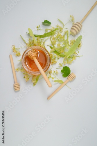 Natural linden honey in a clear glass jar with a wooden honey dipper surrounded by fresh linden blossoms. Minimal food composition on a white background.