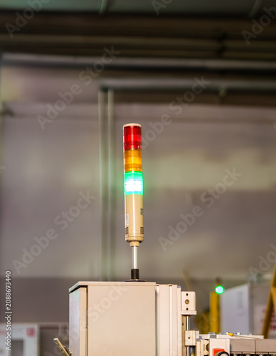 Bild auf Leinwand A glowing industrial signal tower light with red, yellow, and green indicators stands mounted atop a machinery control box within a manufacturing facility