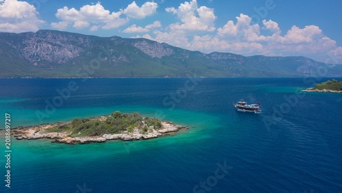 A small island off the coast of Sedir Adası. A tour boat heading towards Akyaka,  lush green mountains rise under a white cloudy sky on the background. Aerial view. Gulf of Gökova, Muğla, Turkey