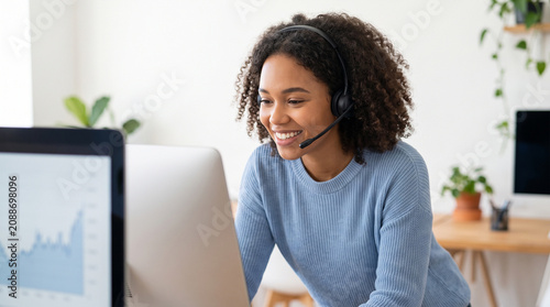 A cheerful young female customer service representative wearing a wireless headset smiles warmly at her computer screen while working remotely in a bright minimalist office.