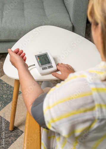 Rear view of middle aged woman checking her blood pressure while sitting on sofa at home. Home healthcare, self monitoring, heart health and preventive medical care.