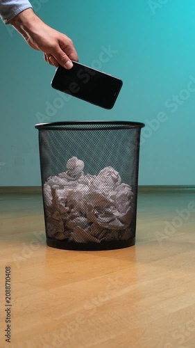 Vertical video human hand dropping an old smartphone into a black mesh trash can filled with crumpled paper. Symbolizing digital detox. Electronic waste