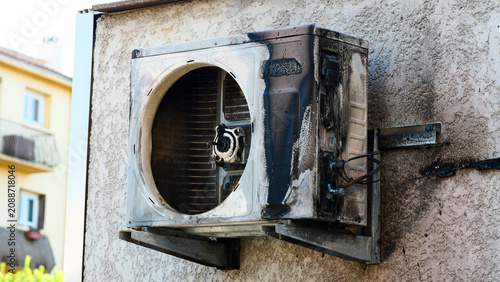 Burnt air conditioner outdoor unit melted from fire or extreme heatwave. Exploded AC system split block damage on house wall due to electricity overload and abnormal summer hot weather climate.