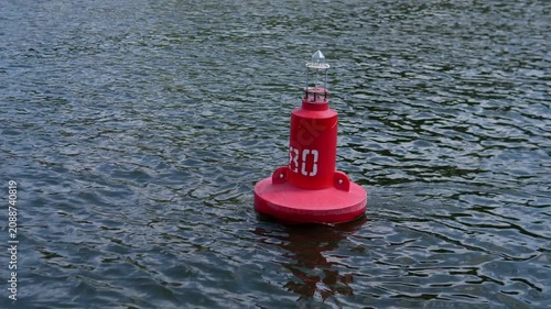 Bright red navigational buoy floats on rippled open water, positioned slightly right of center in foreground from elevated viewpoint, with white number marking, small metal top