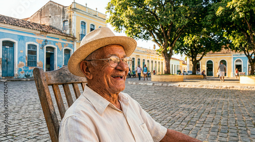Idoso brasileiro rindo em enquadramento grande angular, usando chapéu de palha, sentado em praça de paralelepípedos com casario colonial