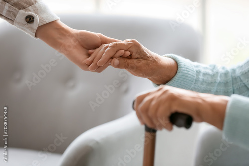 Senior woman with stick and daughter holding hands at home, closeup