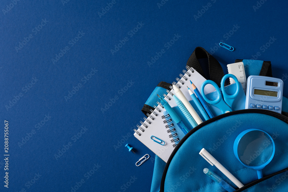 Fototapeta premium Back to school backpack with blue stationery, notebook, pencil, scissors and calculator arranged on blue background