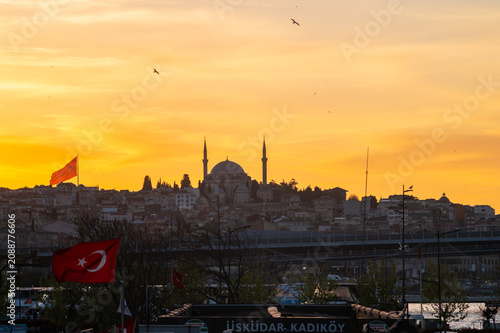 Horizontal photo of a city landscape with a yellow orange sunset with birds , cloudy sky, urban buildings and a mosque in the center.