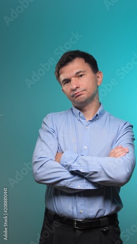 Vertical video man looking at the camera with a skeptical or thoughtful expression, crossing his arms and standing against a vibrant turquoise blue background with ample negative space