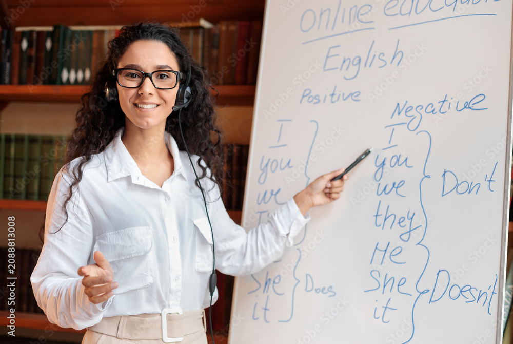 Fototapeta premium Positive young school teacher working from home, teaching English language grammar rules subject to students online. Woman pointing at whiteboard, talking with headphone.