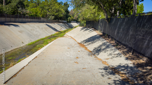 Photography A concrete storm drain channel or urban river bed runs beneath green trees and a