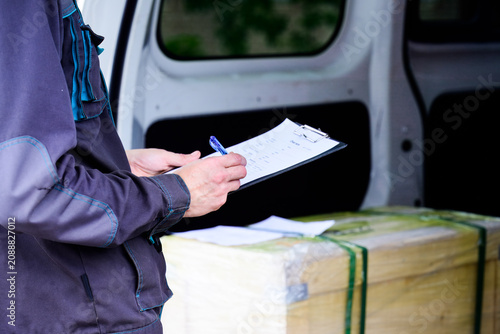 Worker hand writing on clipboard invoice document near wooden crate delivery box in open back cargo van for shipping