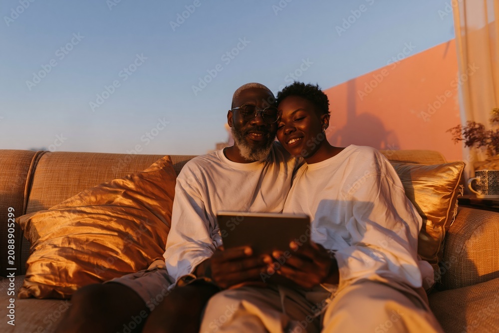 Obraz premium Couple sitting on a sofa using a digital tablet at sunset, glowing golden light on a warm terrace for home technology and leisure visuals