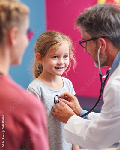Friendly pediatric doctor examining a smiling child using a stethoscope inside a colorful modern examination room, caring parents nearby, authentic healthcare interaction, natural emotions