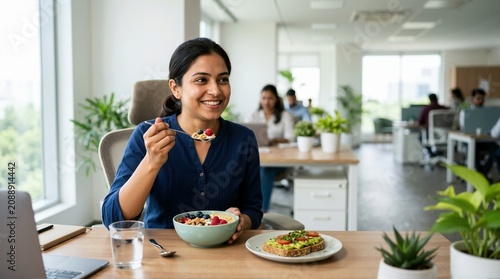 Smiling South Asian businesswoman eating healthy breakfast at office desk. Nutritious meal with yogurt bowl and avocado toast during work break