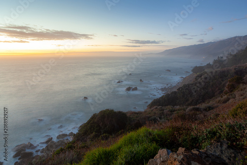 The Pacific Ocean meets the rugged coastline of Big Sur, California, at dusk. The soft light of sunset casts a serene glow over the misty waters and rocky shores.